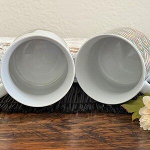 May include: Two white ceramic mugs with a handle. One mug has a colorful geometric pattern. The mugs are placed on a dark surface with a white log and a flower in the background. The mugs are empty.