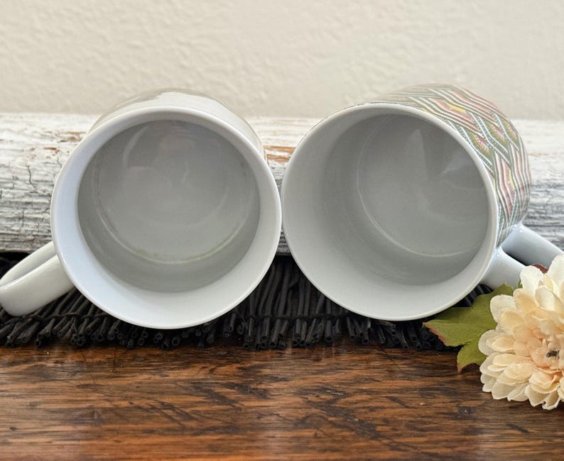 May include: Two white ceramic mugs with a handle. One mug has a colorful geometric pattern. The mugs are placed on a dark surface with a white log and a flower in the background. The mugs are empty.