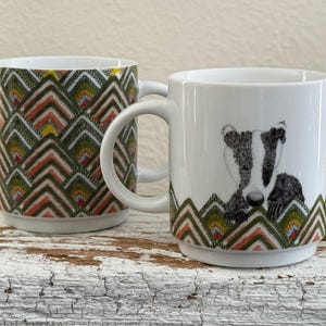 May include: Two white ceramic mugs. One mug features a repeating geometric pattern in shades of green, orange, and yellow. The other mug has a badger illustration in black and white, with a matching geometric pattern at the base.