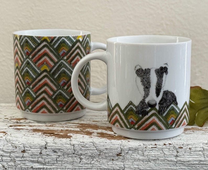 May include: Two white ceramic mugs. One mug features a repeating geometric pattern in shades of green, orange, and yellow. The other mug has a badger illustration in black and white, with a matching geometric pattern at the base.