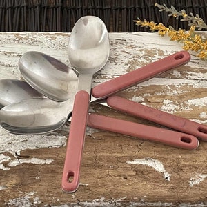 May include: A set of stainless steel spoons with rose-colored handles. The spoons are arranged on a distressed, white-painted wooden surface. The handles have a hole at the end for hanging.