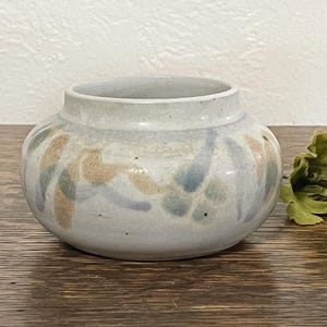 May include: A small, round, ceramic vase with a wide opening. The vase is light gray with a pattern of blue, tan, and green leaf-like shapes around the body. The vase sits on a wooden surface.