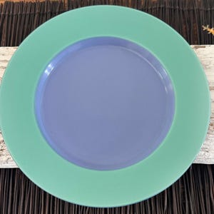May include: A round, ceramic plate with a green outer rim and a light blue center. The plate is a solid color with no patterns or designs.