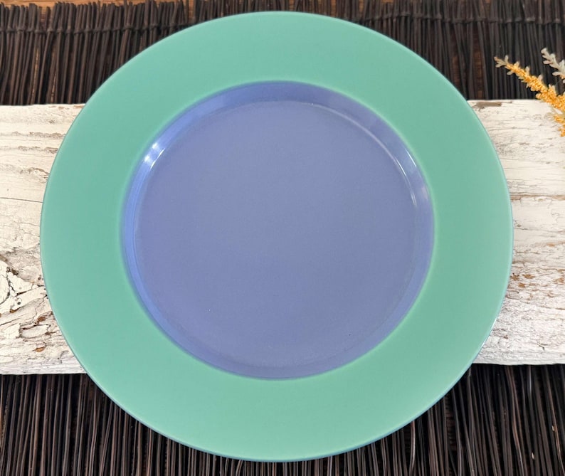 May include: A round, ceramic plate with a green outer rim and a light blue center. The plate is a solid color with no patterns or designs.