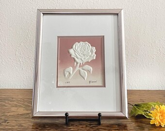 May include: Framed artwork featuring a white rose in relief against a pink and beige gradient background. The silver-toned frame surrounds the artwork, which is displayed on a wooden surface. The artwork is signed and numbered 1/875.