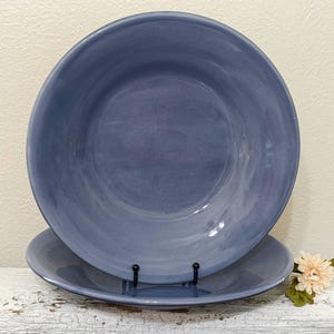May include: Two blue ceramic bowls, one larger than the other, are displayed on a white, distressed wooden surface. The bowls have a matte finish and a gradient of blue hues. A small, decorative flower is placed to the side.