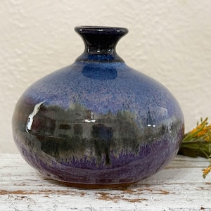 May include: A small, round ceramic vase with a wide mouth and a narrow neck. The vase is glazed in shades of purple, blue, and black, with a mottled effect.