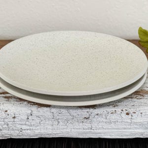 May include: Two off-white ceramic plates stacked on top of each other. The plates have a speckled texture and a slightly raised rim. The plates are on a distressed white wooden surface.