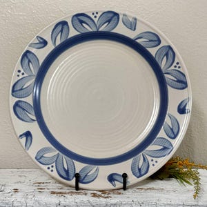 May include: A white ceramic dinner plate with a blue floral design around the rim. The plate has a slightly raised center and a smooth, glazed surface.