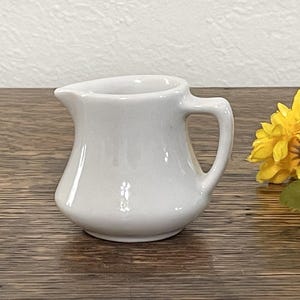 May include: A small, white ceramic creamer with a rounded body and a handle. The creamer is sitting on a wooden surface.