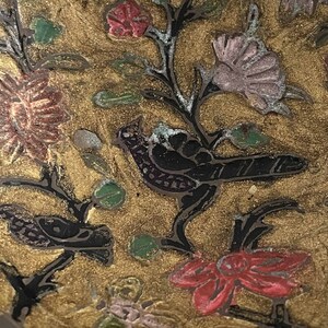 以下が含まれることがあります： 金色の背景に、赤、ピンク、緑の色合いの複雑な花柄が施された装飾的なボウルをクローズアップで撮影したものです。ボウルには、複雑なディテールが施された黒い鳥が2羽描かれています。