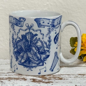 May include: White ceramic mug with a blue musical instrument design. The design includes a violin, trumpet, harp, and musical notes. The mug has a curved handle and is set against a rustic, white wooden surface.