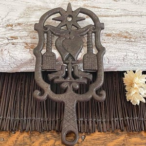 May include: A dark brown cast iron trivet featuring a central heart motif flanked by two vertical tools and two birds at the base. The top is adorned with a star shape, and the handle has a textured diamond pattern with a circular hole at the end.