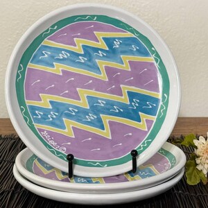May include: Set of four white ceramic plates with a hand-painted design. Each plate features a central pattern of purple, yellow, and teal zigzags, framed by a green border and a white rim. The plates are stacked on a black metal stand.