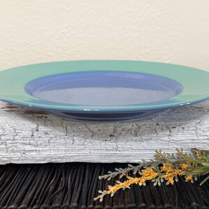 May include: A blue and green ceramic plate with a raised rim. The plate is on a white, weathered surface.