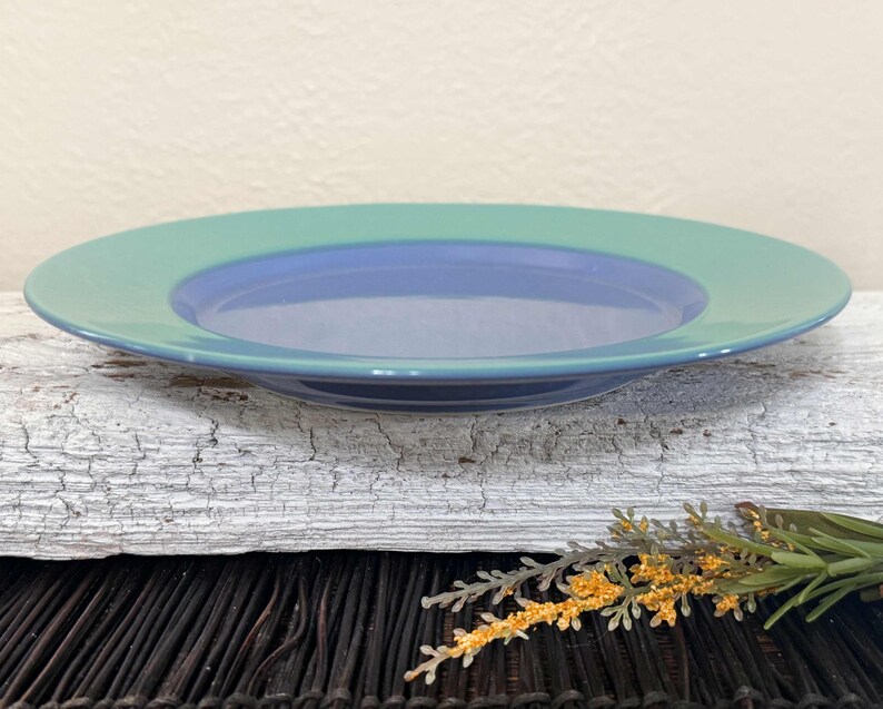 May include: A blue and green ceramic plate with a raised rim. The plate is on a white, weathered surface.