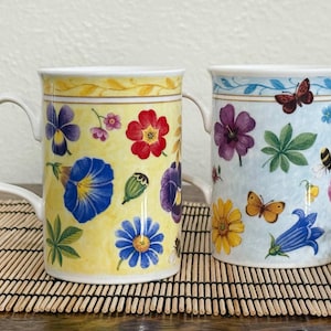 May include: Two ceramic mugs featuring botanical illustrations. The left mug has a yellow background with red, blue, and purple flowers. The right mug has a light blue background with butterflies, a bee, and various colorful floral patterns.