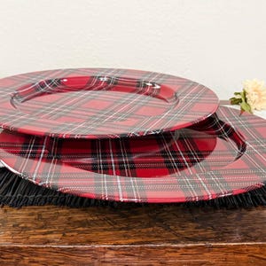 May include: Two round dinner plates featuring a red, black, and white plaid pattern. The plates have a smooth, glossy finish with a wide rim and a central recessed area. The tartan design consists of intersecting lines of varying thickness.