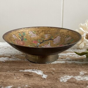 以下が含まれることがあります： 金色に輝く台座と、ピンク、グリーン、レッドの花柄が施された、小さな丸い真鍮製のボウル。ボウルは木製の表面の上に置かれています。