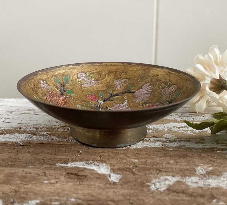 以下が含まれることがあります： 金色に輝く台座と、ピンク、グリーン、レッドの花柄が施された、小さな丸い真鍮製のボウル。ボウルは木製の表面の上に置かれています。