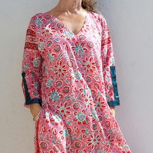 May include: A pink floral print dress with a V-neck and long sleeves. The dress features a red and teal floral pattern and teal trim on the sleeves. The dress is ankle-length and has a relaxed fit.