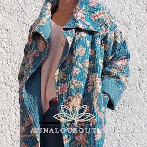 May include: A long, teal quilted coat featuring a floral pattern in shades of pink, red, and cream. The garment has a wide collar, long sleeves, and side pockets. A logo with a lotus icon and the text ANNALOUBOUTIQUE is centred on the coat.