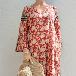 May include: A red maxi dress with a floral print and black trim. The dress has a V-neckline and 3/4 sleeves. The model is wearing a straw hat and sandals.