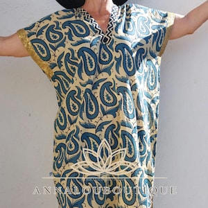 Peut inclure: Un caftan couleur crème avec un imprimé cachemire bleu, un col en V bordé d'un motif chevron noir et blanc, et des revers de manches dorés. Le vêtement présente un motif de lotus blanc central et le texte ANNALOUBOUTIQUE en lettres blanches.