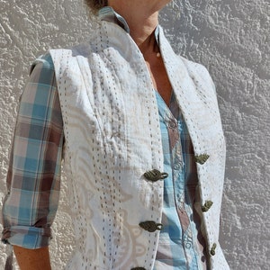 Peut inclure: Un gilet matelassé blanc avec un col montant et des coutures décoratives. Le gilet a quatre fermetures à boutons marrons et un motif floral subtil.