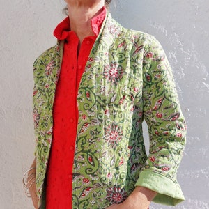 Può includere: Una giacca trapuntata verde con stampa floreale. La giacca ha un colletto alla coreana e maniche lunghe. La giacca è indossata sopra una camicia rossa e jeans blu.