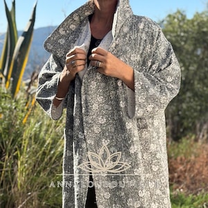 May include: A long, open-front coat featuring a grey and white floral block print pattern. The garment has a wide collar, three-quarter length sleeves, and a white interior lining. The text ANNALOUBOUTIQUE is printed in white with a lotus icon above it.
