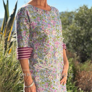 Op de afbeelding: Een lichtblauwe tuniek met een herhalend bloemenpatroon in roze en witte tinten. Het kledingstuk heeft driekwartmouwen met donkerpaarse en witte gestreepte boorden, een ronde halslijn en zijsplitten aan de zoom. Tekst luidt ANNALOUBOUTIQUE.