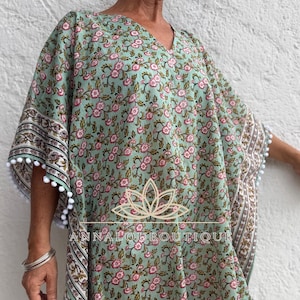 Peut inclure: Un caftan vert clair orné d'un motif répétitif de petites fleurs roses et de feuilles vert foncé. Le vêtement présente des manches larges bordées de pompons blancs et une bordure imprimée à l'ourlet. Un logo lotus doré et le texte ANNALOUBOUTIQUE sont visibles.