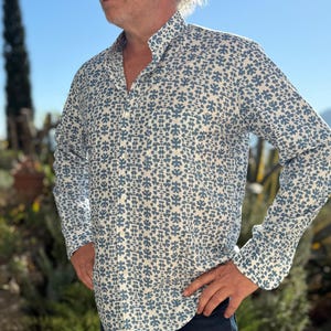 Peut inclure: Chemise à manches longues avec un motif floral bleu sur fond blanc. La chemise a un col classique et est associée à un pantalon bleu foncé. La photo est prise en extérieur, à la lumière naturelle.