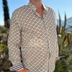 Peut inclure: Une chemise à manches longues boutonnée avec un motif floral répétitif bleu et beige sur fond crème. La chemise possède un col officier et des poignets avec une doublure rayée bleue et blanche. Le texte ANNALOUBOUTIQUE est imprimé sur le devant.