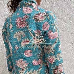 May include: A long-sleeve quilted jacket in a vibrant teal blue fabric featuring a repeating floral pattern in shades of pink, white, and brown. The garment displays visible white running stitch details throughout and includes the text ANNALOUBOUTIQUE.