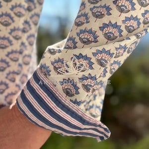 May include: A long-sleeved shirt featuring a repeating blue and grey floral pattern on an off-white background. The cuff is rolled up to reveal a contrasting interior lining with thin blue and red stripes. The text ANNALOUBOUTIQUE is visible.