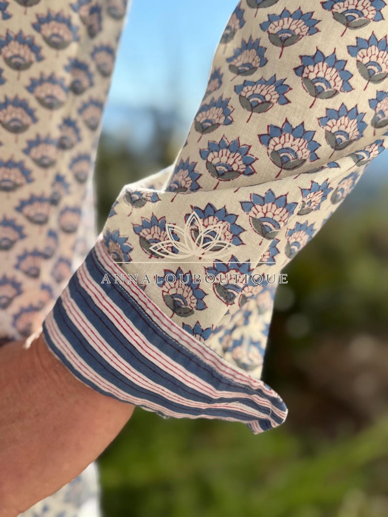 May include: A long-sleeved shirt featuring a repeating blue and grey floral pattern on an off-white background. The cuff is rolled up to reveal a contrasting interior lining with thin blue and red stripes. The text ANNALOUBOUTIQUE is visible.