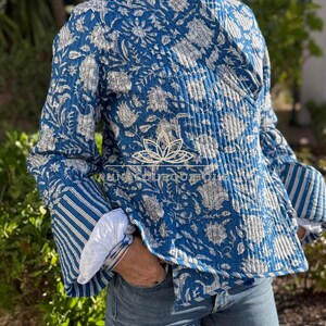 Könnte beinhalten: Eine gesteppte Wickeljacke in Blau und Weiß mit floralem Blockdruckmuster, langen Ärmeln mit gestreiften Bündchen und einem V-Ausschnitt. Das Kleidungsstück zeigt ein Logo mit einer Lotusblüten-Ikone und dem Text ANNALOUBOUTIQUE auf der Vorderseite.