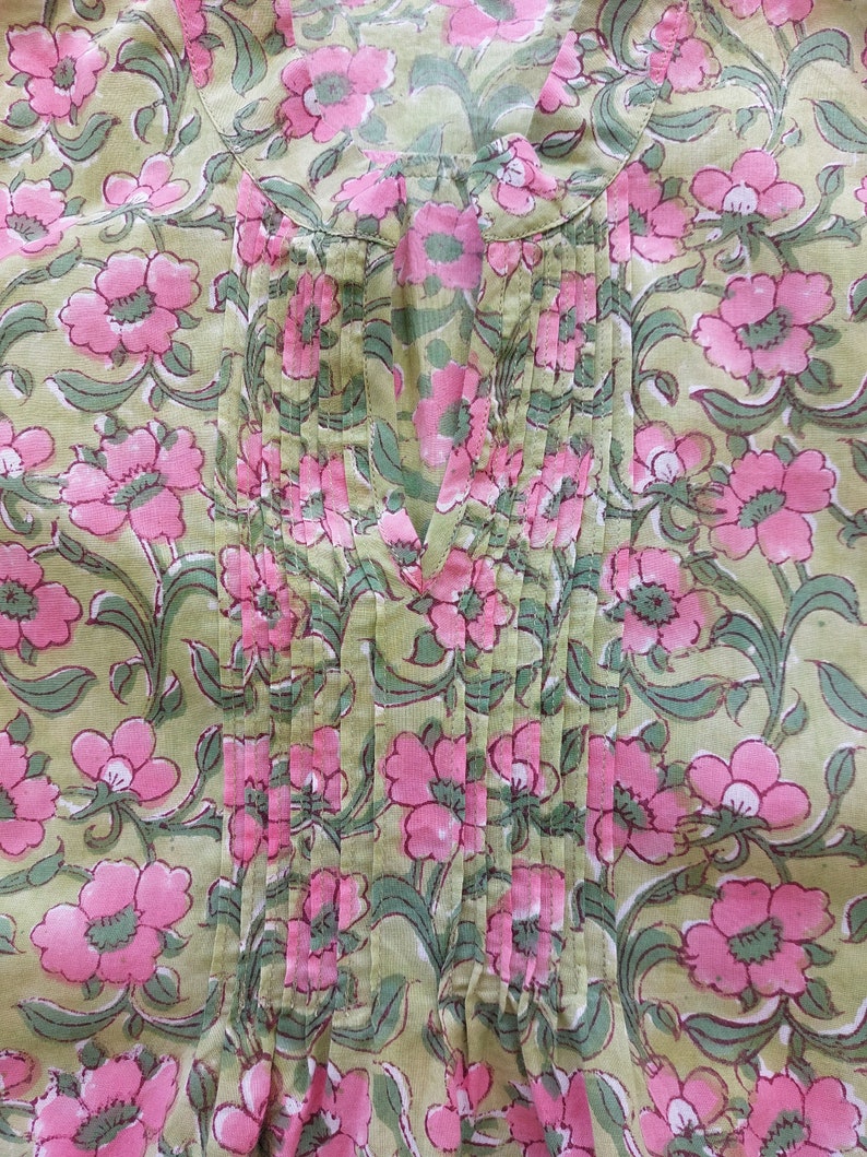 May include: A light green tunic featuring a repeating floral pattern of pink blossoms with dark green stems and leaves. The garment includes a vertical pleated panel down the center front, a notched neckline, and a mandarin-style collar detail.