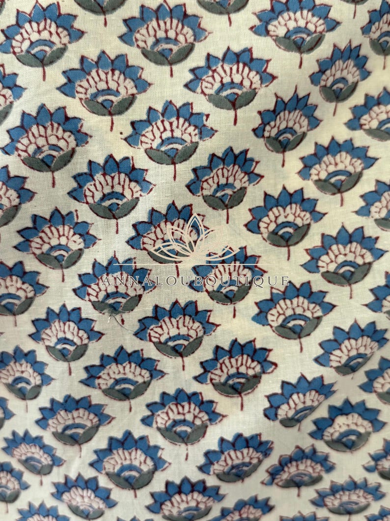 May include: A cream-colored fabric featuring a repeating pattern of blue and dark red lotus-style flowers. Centered on the textile is a white graphic logo of a lotus flower above the text ANNALOUBOUTIQUE in a clean, sans-serif font.