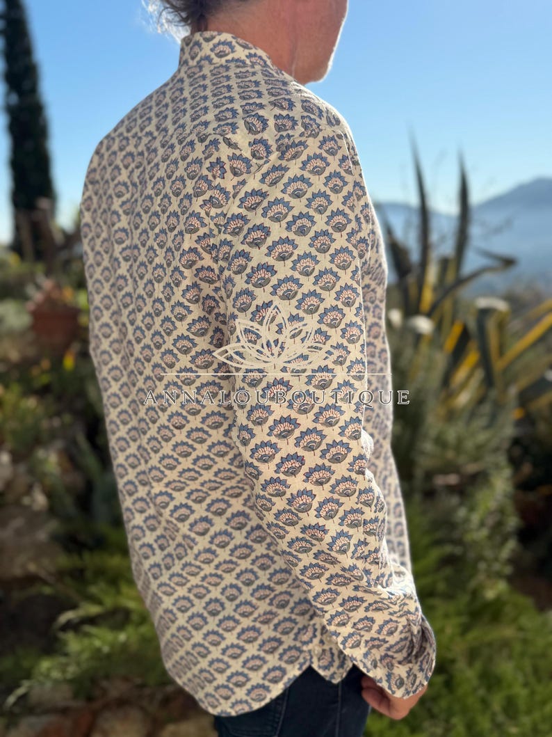 May include: A long-sleeved button-down shirt in an off-white fabric featuring a repeating blue and brown floral block print pattern. The shirt displays a collar and a watermark logo that reads ANNALOUBOUTIQUE with a lotus flower icon.