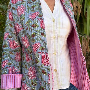 Könnte beinhalten: Hellblaue Steppjacke mit einem Blumenmuster in Rosa und Grün. Die Jacke hat ein rosa-weiß gestreiftes Futter und Manschetten. Sie wird über einem weißen Hemd mit Knöpfen getragen.