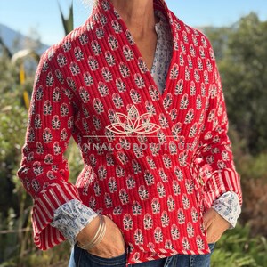 Könnte beinhalten: Eine rote, gesteppte Wickeljacke mit einem sich wiederholenden dunkelblauen und weißen Blumenmuster, langen Ärmeln mit gestreiften Bündchen und einem Schalkragen. Der Text ANNALOUBOUTIQUE ist in Weiß auf der Jacke aufgedruckt.
