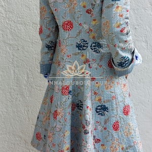 May include: A light blue quilted coat featuring a vibrant floral pattern with red, dark blue, and yellow blossoms. The garment has long sleeves with folded cuffs, a flared hem, and a logo with the text ANNALOUBOUTIQUE printed on the back.