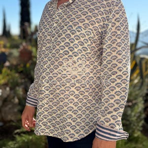 May include: A long-sleeved button-down shirt featuring a repeating blue and tan floral pattern on a cream background. The cuffs display a blue and white striped design. The text ANNALOUBOUTIQUE is printed across the center of the shirt.