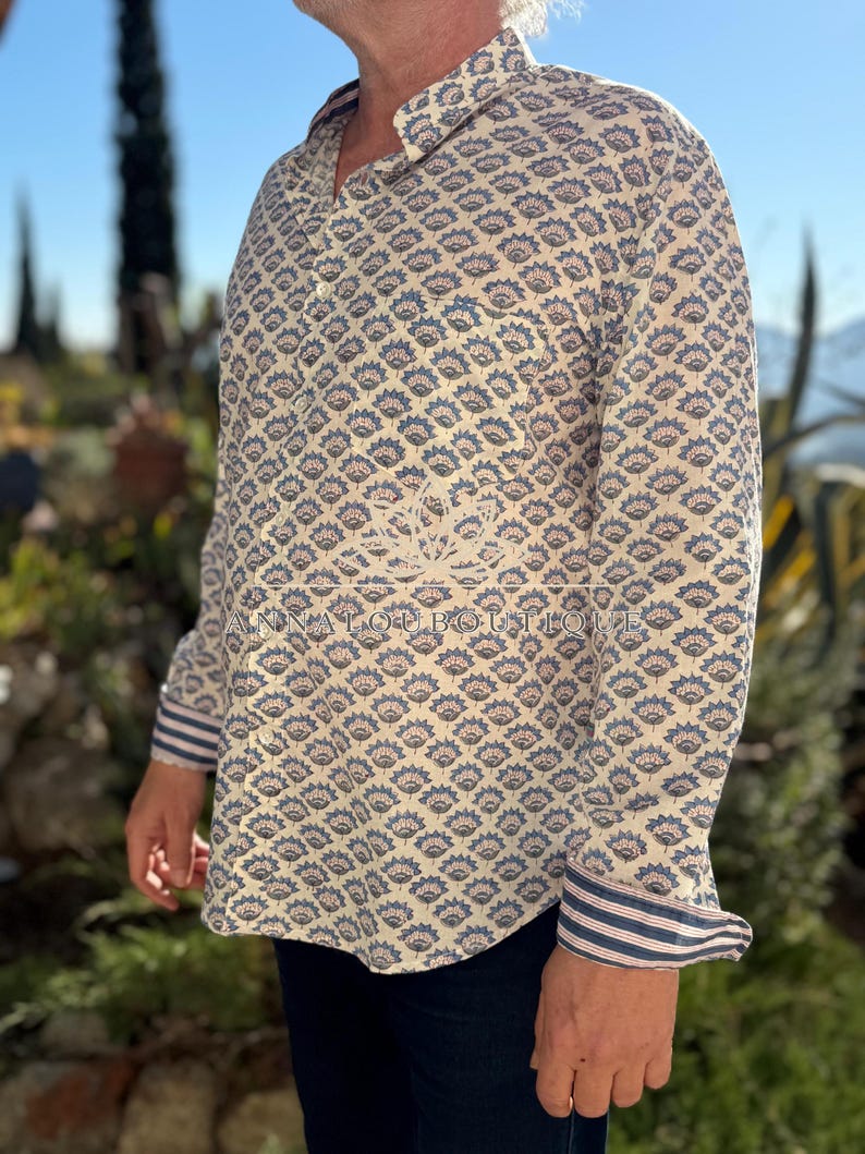 May include: A long-sleeved button-down shirt featuring a repeating blue and tan floral pattern on a cream background. The cuffs display a blue and white striped design. The text ANNALOUBOUTIQUE is printed across the center of the shirt.