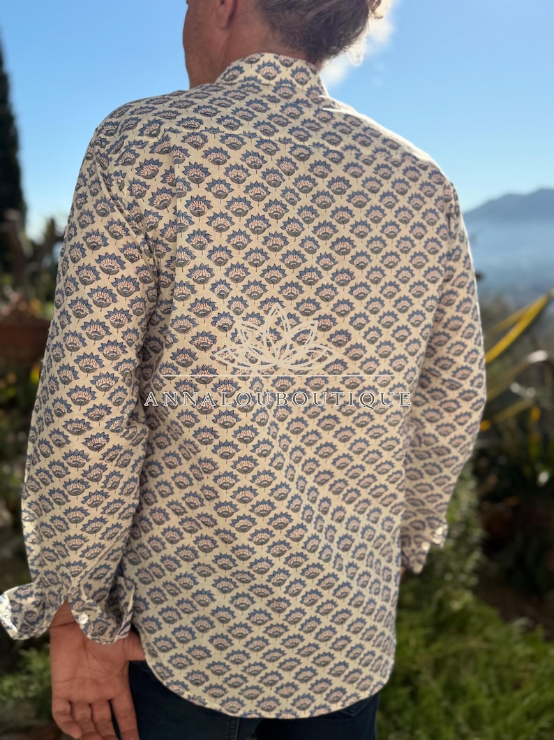 May include: A long-sleeve button-down shirt in a cream color featuring a repeating blue and orange floral pattern. The back of the shirt displays a white lotus flower graphic and the text ANNALOUBOUTIQUE printed across the center.