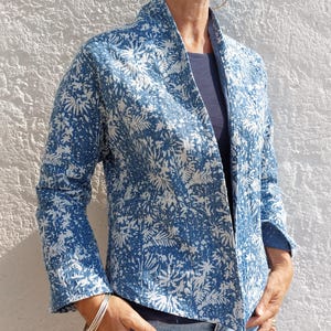 Könnte beinhalten: Eine blau-weiße Steppjacke mit botanischem Blatt- und Blumenmuster, Schalkragen und langen Ärmeln. Das Kleidungsstück ist mit sichtbaren Nähten durch den gesamten Stoff gefertigt, was eine strukturierte Optik mit einer offenen Vorderseite erzeugt.