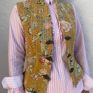Peut inclure: Gilet matelassé moutarde à motifs floraux roses, verts et blancs. Le gilet est orné de fermetures en forme de feuilles vert foncé et porté sur une chemise rayée rose et blanche avec des volants.